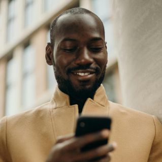 A cheerful man in a stylish coat using his smartphone and holding coffee outside.