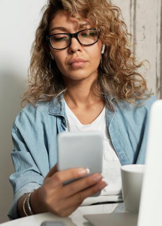 Woman using smartphone and laptop while working remotely in a home kitchen setting.