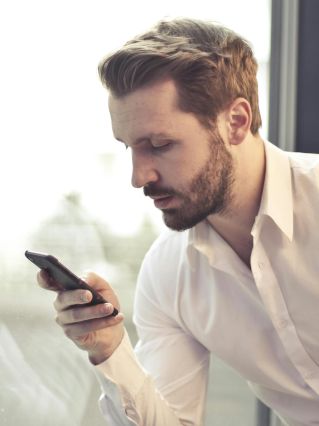 A businessman in a white shirt texts on his smartphone by a large glass window inside an office.